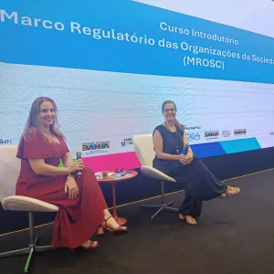 Em um palco, duas mulheres estão sentadas em poltronas brancas, lado a lado, cada uma segurando um microfone. À esquerda, uma mulher de vestido vermelho longo sorri para a câmera. À direita, outra mulher veste roupa escura e também sorri. Entre elas, há uma pequena mesa com objetos. Ao fundo, um telão grande exibe o título “Curso Introdutório – Marco Regulatório das Organizações da Sociedade Civil (MROSC)” e logotipos de instituições. O ambiente parece ser um evento ou palestra em Salvador.