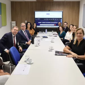 Sala de reunião com mesa longa e pessoas sentadas ao redor. Homens e mulheres, em trajes formais, participam de uma conversa. Alguns anotam, outros escutam com atenção. Ao fundo, uma tela exibe o título “Semana de Saneamento, Precedentes e Ações Coletivas”. O ambiente é claro, com paredes neutras e painéis coloridos.