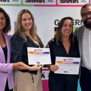 Quatro pessoas estão lado a lado, em pé, sorrindo para a foto. Ao fundo, há um painel colorido com o logotipo do Governo do Estado da Bahia repetido. Duas mulheres no centro seguram certificados com as duas mãos. À esquerda, uma mulher de blazer lilás apoia o certificado de uma delas. À direita, um homem de barba, óculos e blazer cinza coloca o braço em volta do grupo. Todos usam roupas formais. A cena transmite um momento de entrega de certificados.
