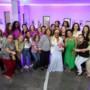 Grupo de mulheres posa para foto em um espaço interno com iluminação roxa. Elas estão lado a lado e também agachadas na frente, sorrindo e olhando para a câmera. No centro, duas mulheres se destacam: uma de blusa cinza com bolinhas e outra com vestido claro e lenço na cabeça, segurando um pequeno presente. As roupas são variadas, com cores como rosa, verde, preto e estampas. Ao fundo, há colunas, quadros na parede e luzes decorativas. O clima é alegre e de celebração.