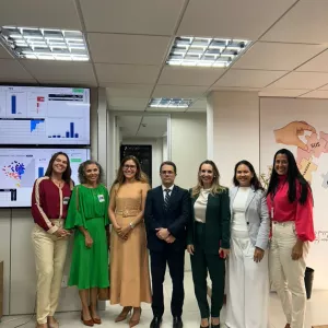 Grupo de sete pessoas posa para foto em um ambiente institucional. Ao fundo, há telas exibindo painéis com gráficos e dados. Os participantes estão alinhados lado a lado, vestidos com roupas formais e crachás de identificação, em frente a um espaço de monitoramento. O ambiente é bem iluminado e organizado, sugerindo uma apresentação ou visita técnica.