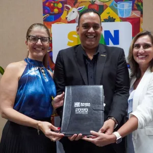 Três pessoas posam sorrindo para a foto em um ambiente interno. Ao centro, um homem de terno preto segura uma pasta preta com as mãos. A pasta tem o texto “Bahia Sem Fome” e “Governo do Estado Bahia”. À esquerda, uma mulher de óculos usa blusa azul sem mangas e segura a pasta junto com ele. À direita, outra mulher, com blazer branco, também segura a pasta. Ao fundo, há um painel colorido com ilustrações de alimentos e uma planta à esquerda.