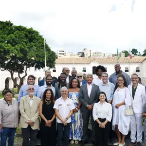 Grupo de cerca de vinte pessoas posa para uma foto ao ar livre. Elas estão organizadas em duas fileiras, em pé, sobre um pequeno palco. A maioria veste roupas sociais, como ternos, camisas e vestidos. Ao fundo há um prédio histórico de dois andares, com paredes claras, janelas verdes e telhado de telhas claras. Bandeiras estão hasteadas no centro do edifício. À esquerda há uma grande árvore com copa cheia e, ao redor, um jardim com gramado. Algumas pessoas aparecem ao fundo caminhando pelo espaço.