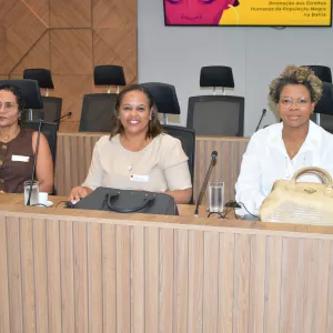 Três mulheres estão sentadas lado a lado em uma mesa de reunião, olhando para a câmera. Sobre a mesa há bolsas, copos de água e equipamentos de áudio. Ao fundo aparecem cadeiras e um painel de madeira na parede.