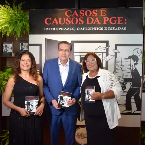 Uma foto em grupo com três pessoas de pé e sorrindo, segurando exemplares de um livro. Da esquerda para a direita, temos uma mulher de vestido preto longo de um ombro só, um homem de terno azul escuro e uma mulher de blazer branco e blusa preta. O fundo é composto por um painel preto com texto e ilustrações em branco, prateleiras de madeira com livros e vasos de plantas, e o logotipo da PGE com a inscrição "PROCURADORIA GERAL DO ESTADO DA BAHIA" e "60 ANOS".