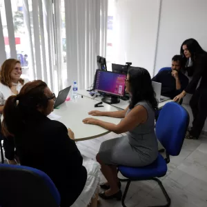 Um grupo de cinco pessoas está reunido em um escritório ao redor de uma mesa redonda. Duas mulheres conversam sentadas, enquanto outra trabalha em um notebook. Ao fundo, um homem está sentado em frente ao computador e uma mulher está em pé ao lado dele. O ambiente é claro, com cadeiras azuis, mesas e equipamentos de trabalho.