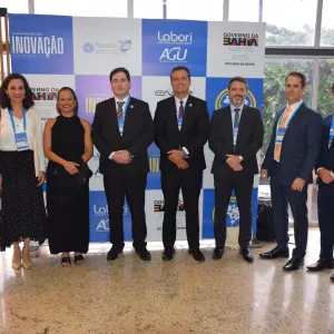 Grupo de pessoas posando para foto em frente ao painel do evento “Caravanas de Inovação”. Todos estão alinhados, vestidos formalmente e com crachás. O ambiente é iluminado, com janelas ao fundo mostrando a área externa.