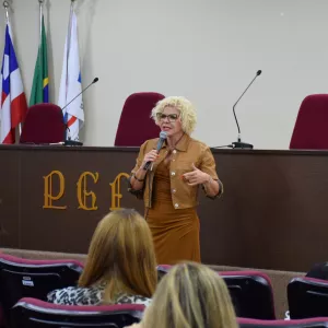 Mulher de cabelo curto e cacheado, loiro claro, está em pé à frente de um auditório, segurando um microfone. Usa óculos e roupa em tom marrom. Ela gesticula com uma das mãos enquanto fala, demonstrando expressão séria e envolvida. Ao fundo, há uma mesa com cadeiras e três bandeiras. Na frente dela, pessoas estão sentadas em cadeiras vermelhas, vistas de costas, assistindo à apresentação. O ambiente é organizado e lembra uma palestra ou evento institucional.