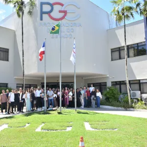 Foto externa, em dia ensolarado. Ao centro, um prédio branco com a sigla “PGE” e o nome “Procuradoria Geral do Estado” na fachada. Em frente, três mastros com bandeiras, incluindo a do Brasil. No gramado, as letras “PGE” formadas com pedras. Um grande grupo de pessoas está reunido na entrada, posando para a foto. Há palmeiras altas ao redor e janelas nas laterais do prédio. A cena transmite um encontro institucional.