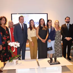 Grupo de várias pessoas alinhadas para uma foto em evento formal. Homens e mulheres vestem roupas sociais e estão sorrindo. Ao fundo, há um telão com logotipo e decoração simples, indicando uma cerimônia ou encontro institucional.