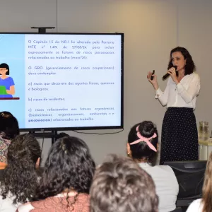 Uma mulher apresenta conteúdo exibido na tela, segurando um microfone. O slide mostra informações sobre normas e riscos no trabalho. Parte do público aparece de costas, acompanhando atentamente a explicação.