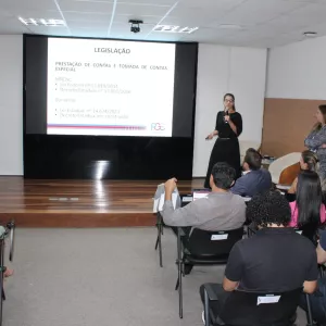 Uma palestrante de vestido preto fala ao microfone no palco, apontando para um telão que detalha informações sobre "LEGISLAÇÃO" e "PRESTAÇÃO DE CONTAS". Dois outros palestrantes acompanham a fala nas laterais do palco.
