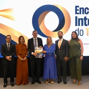 Seis pessoas estão em pé, lado a lado, diante de um grande painel iluminado. Ao fundo, aparece a imagem de um farol, um símbolo circular em azul e laranja e o texto “Encontro de Integridade do TJBA”. As pessoas usam roupas sociais; duas vestem ternos, três usam vestidos e uma usa blusa e calça. A pessoa ao centro segura um certificado ou material do evento. Todos posam para a foto.