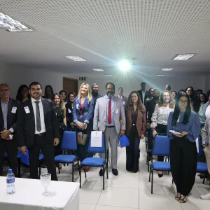 Fotografia de um grupo numeroso de pessoas reunidas em uma sala. Elas estão em pé e voltadas para a câmera, muitas sorrindo. A maioria usa roupas sociais, como ternos, blazers, camisas e vestidos, e algumas usam crachás. Ao centro, há um homem de terno cinza e gravata vermelha. À frente, há cadeiras azuis. No canto inferior esquerdo, uma mesa coberta com toalha branca exibe garrafas de água e taças. O ambiente tem teto com iluminação embutida e ar-condicionado.