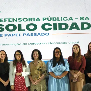 Em frente a um painel com o texto “Defensoria Pública – BA” e “Solo Cidadão – De Papel Passado”, sete pessoas posam para uma foto. Da esquerda para a direita, há três homens e quatro mulheres, vestidos formalmente. As mulheres usam roupas em preto, branco, vermelho, bege e azul-claro. Todos estão de pé, lado a lado, sorrindo ou olhando para a câmera. O painel também traz a frase “Apresentação de Defesa da Identidade Visual”.