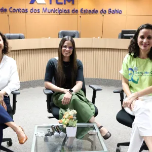 Fotografia de três mulheres sentadas em cadeiras, em um ambiente institucional. A mulher à esquerda usa camisa branca, calça azul-marinho e sapatos marrons. A mulher ao centro veste blusa preta, calça verde, óculos e relógio. A mulher à direita usa camiseta verde-clara com estampa, calça branca e tênis. Todas sorriem para a câmera. À frente delas, há uma mesa de vidro com microfones e um pequeno arranjo de flores. Ao fundo, vê-se um painel de madeira com a identificação do TCE-BA