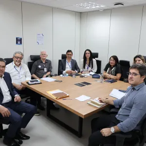 Sala de reunião com paredes claras, mesa retangular de madeira e cadeiras pretas. Nove pessoas estão sentadas ao redor da mesa, voltadas para a câmera. Algumas usam crachás e estão com papéis, cadernos, celulares e canetas sobre a mesa. Ao fundo, há uma tela de televisão, cartazes nas paredes e luminárias no teto.