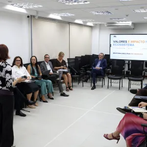 Fotografia de uma reunião em uma sala ampla e clara, com pessoas sentadas em cadeiras dispostas em semicírculo. Ao centro, uma mulher está em pé, de costas para a câmera, conduzindo a atividade. Os participantes fazem anotações e utilizam computadores. Ao fundo, há uma tela exibindo a apresentação “Valores e Impacto no Ecossistema” e informações sobre mentoria executiva em liderança.