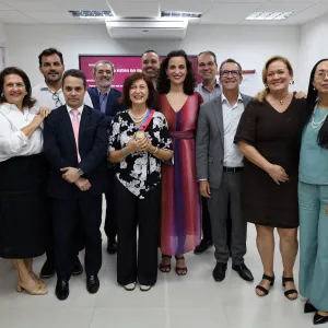 Fotografia de um grupo de onze pessoas reunidas em uma sala. Elas estão em pé, lado a lado, sorrindo e olhando para a câmera. Ao centro, uma mulher usa blusa preta com estampa de flores brancas, colar de pérolas e segura uma medalha. Os demais participantes usam roupas sociais e casuais. Ao fundo, há uma tela com uma apresentação e cadeiras organizadas pela sala.