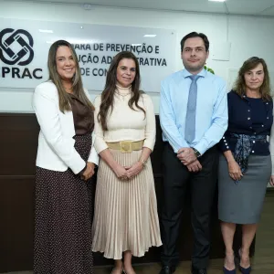 Foto de cinco pessoas em pé, lado a lado, em um ambiente institucional. Ao fundo, há um balcão de madeira e uma placa com o logotipo e a sigla “CPRAC”. Da esquerda para a direita, estão duas mulheres, um homem e duas mulheres. As mulheres usam roupas sociais, com blazers, saias e vestidos. O homem veste camisa azul-clara, gravata estampada e calça escura. Todos olham para a câmera e sorriem discretamente.
