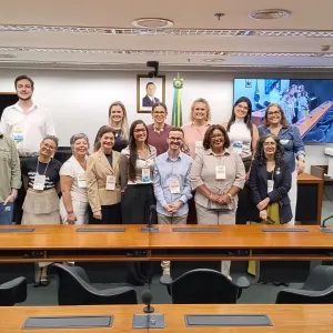 Foto de um grupo de cerca de 14 pessoas reunidas em uma sala de reuniões. Elas estão de pé, lado a lado, atrás de uma longa mesa de madeira com microfones. Ao fundo, há bandeiras, um retrato, um relógio digital e uma tela exibindo outra imagem da sala. Todos posam para a foto e usam crachás de identificação.