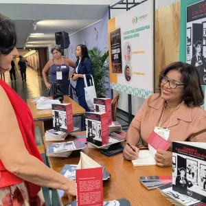 Foto de um estande de evento. À direita, uma mulher de óculos, usando roupa social em tom salmão e crachá, está sentada à mesa, sorrindo e autografando um livro. À esquerda, outra mulher, de costas para a câmera, usa roupa vermelha e carrega uma bolsa da mesma cor. Sobre a mesa, há vários exemplares do livro “Casos e causos da PGE-BA: entre prazos, cafezinhos e risadas”. Ao fundo, outras pessoas conversam e há painéis com materiais de divulgação.