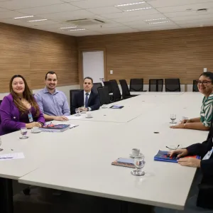 Sala de reuniões ampla, com paredes revestidas de madeira e uma grande mesa branca. Seis pessoas estão sentadas ao redor da mesa, em lados opostos, voltadas para a câmera. Há quatro mulheres e dois homens, todos vestidos de maneira formal ou social. Sobre a mesa, há copos com água, xícaras, celulares, cadernos e documentos. Ao fundo, há cadeiras pretas e uma porta.