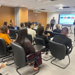 Sala de reunião com várias pessoas sentadas em cadeiras diante de uma tela. Ao fundo, um homem de terno segura um microfone e fala para o grupo. Outro homem está em pé próximo à tela, que exibe uma apresentação. As paredes têm revestimento de madeira, há janelas com persianas, ar-condicionado e projetor no teto. Os participantes observam a apresentação e alguns usam celulares ou computadores.