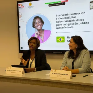 Fotografia de um painel de evento. Duas mulheres estão sentadas à mesa. À esquerda, uma mulher negra, de cabelos curtos, usa blazer preto e blusa clara. À direita, uma mulher de cabelos longos usa blusa cinza. Ao fundo, um telão exibe o título “Boa administração na era digital: Governança de dados para uma gestão pública mais eficiente”, além das imagens e nomes das participantes. Sobre a mesa, há placas de identificação, microfones e objetos pessoais.