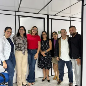 Foto de sete pessoas em pé, lado a lado, em um ambiente com divisórias brancas e estruturas pretas. Da esquerda para a direita, são cinco mulheres e dois homens. As pessoas usam roupas casuais e sociais, em diferentes cores e estampas, e olham para a câmera, sorrindo. Ao fundo, há portas ou divisórias com placas verdes de sinalização. No canto inferior esquerdo, aparecem cadeiras azuis. O piso é claro e o teto tem placas brancas com estrutura preta.