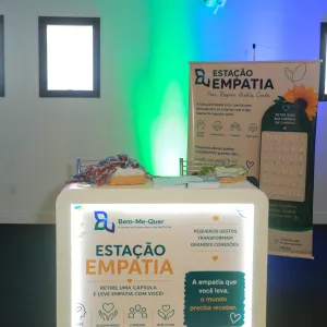 Foto de um espaço de divulgação chamado “Estação Empatia”. Ao centro, há um balcão iluminado, com cartazes sobre empatia, acolhimento, conexão e bem-estar. Sobre o balcão, estão cestas com materiais e pequenos cartões. Ao fundo, um painel vertical traz o mesmo tema, com uma flor, folhas e um suporte com cápsulas de papel. O ambiente é interno, com paredes claras e iluminação decorativa em verde e azul.