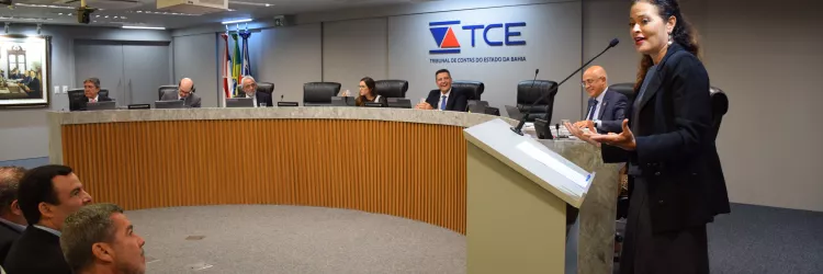 Imagem mostra uma mulher em pé falando ao microfone, posicionada atrás de um púlpito no plenário do Tribunal de Contas do Estado da Bahia (TCE-BA). Ao fundo, sete pessoas estão sentadas em uma bancada semicircular, com laptops e microfones à frente. Todos estão com trajes formais. Três homens sentados na plateia observam a oradora com atenção. Na parede, há o logotipo do TCE e bandeiras ao fundo. O ambiente é bem iluminado, moderno e com clima institucional.