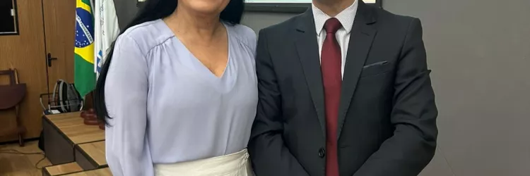 Foto com duas pessoas em pé, sorrindo. À esquerda, mulher branca, cabelos pretos, longos e lisos, usando uma blusa de manga comprida lilás clara e uma saia branca. À direita, homem branco, cabelos curtos e escuros, vestindo terno preto, camisa branca e gravata vermelha. Estão em um ambiente fechado, com piso de madeira clara e uma tela ao fundo escrito: "CERTIFICAÇÃO BIM – Inovação, qualidade e eficiência nas obras do Estado".