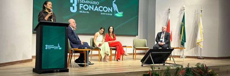 Mulher fala ao microfone em pé, atrás de um púlpito, no palco de um auditório. Ao fundo, uma grande tela exibe “3º Seminário FONACON – Belém-PA – 31/07 e 01/08”. Sentados ao lado da palestrante, há quatro pessoas: três mulheres e dois homens, todos atentos ao evento. À direita do palco, estão três bandeiras: do Pará, do Brasil e uma branca institucional. Na frente do palco, há plantas decorativas com folhagens verdes e flores vermelhas. O ambiente é iluminado, formal e organizado.