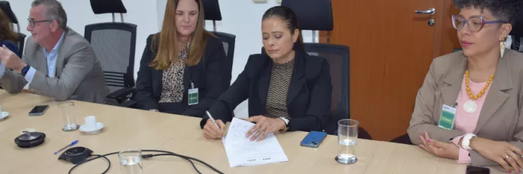 A imagem mostra um grupo de quatro pessoas em uma reunião ao redor de uma mesa. À esquerda, há um homem com cabelo grisalho, usando um terno cinza claro. Ao lado dele, uma mulher com cabelo longo e loiro, vestindo um blazer preto e blusa de estampa. No centro, uma mulher com cabelo preso, usando um terno preto, assina um documento. À direita, uma mulher de cabelo curto e grisalho, com óculos e acessórios coloridos, observa.