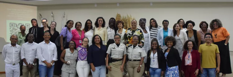 Um grupo diverso de pessoas posa sorrindo para uma foto em um auditório. Elas estão em pé sobre o palco e no chão à frente dele. A maioria são mulheres negras, duas usando uniformes, outras roupas sociais ou casuais. Ao fundo, há bandeiras e um arranjo decorativo com plantas secas. As cadeiras vinho do auditório aparecem na frente da imagem. A foto transmite um clima de encerramento de evento.