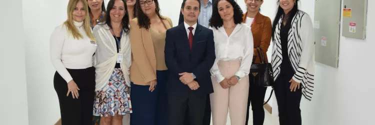Grupo de 10 pessoas, entre homens e mulheres, posando para foto em um corredor de ambiente corporativo. Todos estão sorrindo e vestidos com trajes formais ou sociais.
