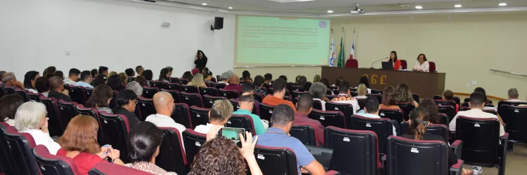 Auditório com várias fileiras de cadeiras ocupadas por pessoas assistindo a uma apresentação. Duas mulheres estão sentadas à mesa no palco, uma delas falando ao microfone. Atrás delas, há quatro bandeiras e uma projeção com texto na parede. O ambiente é iluminado com luzes no teto e tem aparência formal e organizada.