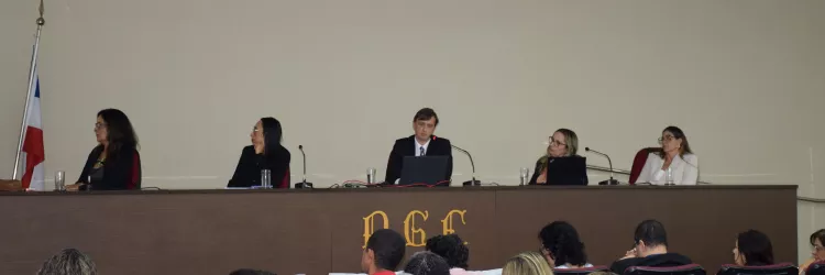 Em uma sala de auditório, várias pessoas estão sentadas assistindo a uma mesa de debate. Ao fundo, cinco pessoas estão sentadas atrás de uma bancada com microfones e laptops. No centro da mesa, um homem de terno fala ao microfone. As outras quatro pessoas, todas mulheres, ouvem atentamente. À esquerda da imagem, há uma bandeira do estado da Bahia. Na parte frontal da bancada está escrita a sigla "PGE". O ambiente é iluminado, e o público está atento à apresentação.