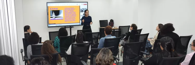 Palestrante apresenta conteúdo sobre Prevenção e Combate em uma sala com várias pessoas sentadas assistindo, olhando para uma tela grande ao fundo com slide colorido.