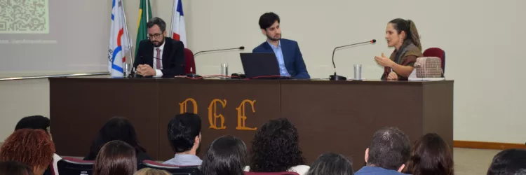 Em um auditório, três pessoas estão sentadas atrás de uma mesa participando de uma mesa-redonda. À esquerda, um homem de terno escuro e gravata vermelha está com as mãos entrelaçadas. No centro, outro homem de terno azul observa a mulher à direita, que fala ao microfone e gesticula com as mãos. Atrás deles, há três bandeiras (Brasil, Bahia e institucional). O público assiste, sentado em cadeiras acolchoadas.