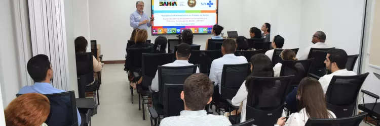 Em uma sala de reuniões clara e moderna, cerca de vinte pessoas estão sentadas, de frente para um palestrante que está em pé ao lado de uma tela. A apresentação exibe o tema “Assistência Farmacêutica no Estado da Bahia”, com logos do Governo da Bahia e do SUS. O público está atento, e a maioria veste roupas sociais. As cadeiras são pretas e a iluminação é branca, vinda do teto. A atmosfera é profissional e de aprendizado.