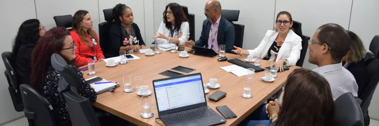 Imagem mostra dez pessoas reunidas em torno de uma mesa de madeira em uma sala de reunião. O grupo é formado por homens e mulheres, todos em trajes sociais. Algumas pessoas falam, enquanto outras escutam com atenção. Sobre a mesa, há copos com água, xícaras de café, celulares, cadernos, teclados e notebooks. Uma das telas do notebook está aberta em um sistema de e-mails. O ambiente é claro, com paredes brancas e cadeiras pretas, transmitindo um clima de reunião formal e colaborativa.