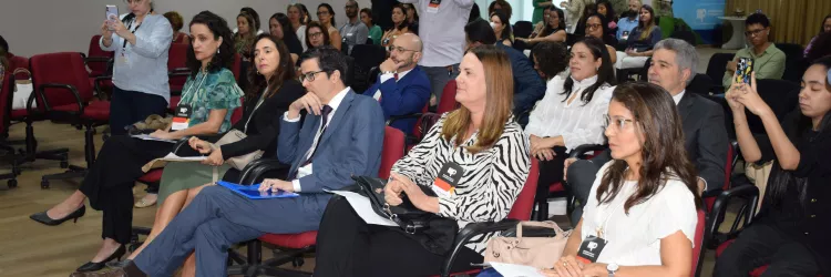 A imagem mostra uma plateia atenta em um auditório. As pessoas estão sentadas em cadeiras vermelhas com rodinhas, e a maioria usa roupas formais. Algumas estão anotando, outras assistindo e algumas tirando fotos com celulares. O ambiente é iluminado, com piso de madeira clara e uma grande imagem do Farol da Barra ao fundo. A cena transmite um clima de evento institucional ou reunião profissional.