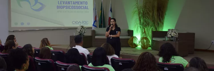 Auditório com público assistindo a uma mulher que está de pé no palco, falando ao microfone. Ela veste roupa preta e está posicionada entre sofás e arranjos decorativos com iluminação verde. Ao fundo, há três bandeiras e um telão exibindo o título da apresentação: “Levantamento Biopsicossocial – Apresentação do Resultado”.