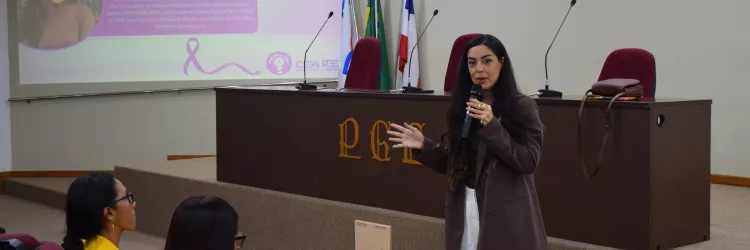 A imagem mostra uma mulher de cabelos longos falando ao microfone em auditório. Ela veste blazer marrom e calça branca. Ao fundo, no telão, aparece a apresentação intitulada “Protocolo de Gênero do CNJ”, com foto e informações da palestrante Daniela Portugal, doutora e mestre em Direito Público pela UFBA. Algumas pessoas sentadas nas poltronas vermelhas acompanham a palestra, enquanto a mesa principal tem bandeiras e microfones.