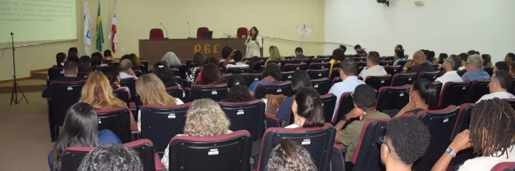 Visão mais ampla do auditório, com a plateia sentada em poltronas vermelhas. A palestrante aparece ao fundo, à frente da tela, explicando o conteúdo jurídico projetado. O local é bem iluminado e há grande participação do público.