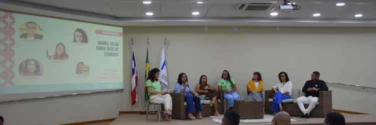Foto de um auditório com um debate em andamento. No palco, sete pessoas estão sentadas em poltronas de vime, participando de uma roda de conversa. Uma das mulheres fala ao microfone. À esquerda, há um telão com o tema do evento: “Vamos falar sobre rede de cuidado?”. No fundo, há três bandeiras: da Bahia, do Brasil e uma branca institucional. O público está sentado em cadeiras vermelhas, assistindo à conversa. O ambiente é iluminado por luzes de teto.