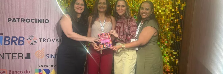 Na imagem, quatro mulheres estão sorrindo e posando juntas em frente a um painel iluminado com a inscrição “51º CNPE”. Elas estão de pé, uma ao lado da outra. A primeira mulher, à esquerda, usa um vestido preto e segura a mão de uma colega. A segunda, de cabelo longo e blusa branca, segura um livro nas mãos. A terceira mulher usa uma blusa vermelha e calça bege. A quarta mulher, à direita, usa um vestido verde e está sorrindo. O fundo é dourado e decorado.