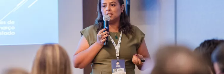 Close da palestrante, mulher de pele clara e cabelos castanhos soltos, usando vestido verde e crachá. Ela segura microfone e controle, enquanto fala ao público atento.